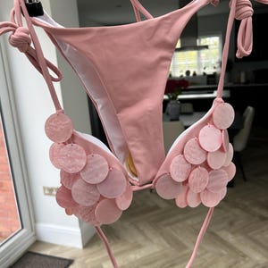 Puede incluir: Un conjunto de bikini rosa claro con lazos. La parte superior presenta adornos circulares rosas decorativos. El bikini est&aacute; colgado de una percha de metal. El fondo es una habitaci&oacute;n con una puerta y una ventana.