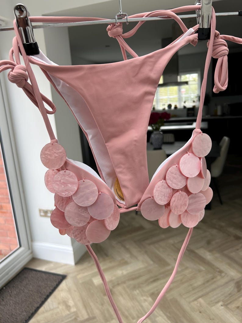 Puede incluir: Un conjunto de bikini rosa claro con lazos. La parte superior presenta adornos circulares rosas decorativos. El bikini est&aacute; colgado de una percha de metal. El fondo es una habitaci&oacute;n con una puerta y una ventana.