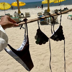 Puede incluir: Un conjunto de bikini marr&oacute;n con un dise&ntilde;o similar a monedas en la parte superior, colgado de un palo de madera. La parte inferior del bikini es marr&oacute;n con un forro blanco. El fondo muestra una playa con sombrillas amarillas y el mar.