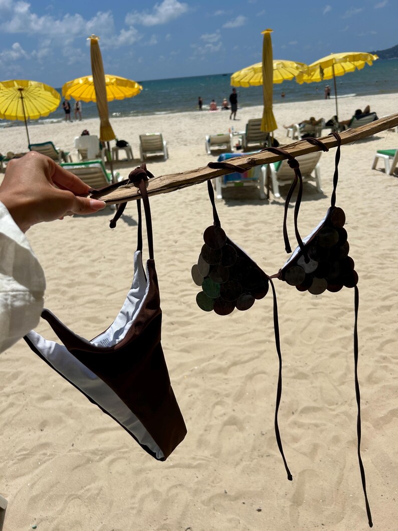 Puede incluir: Un conjunto de bikini marr&oacute;n con un dise&ntilde;o similar a monedas en la parte superior, colgado de un palo de madera. La parte inferior del bikini es marr&oacute;n con un forro blanco. El fondo muestra una playa con sombrillas amarillas y el mar.