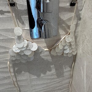 Puede incluir: Un top de bikini de conchas blancas colgado de una percha. El top de bikini est&aacute; hecho de peque&ntilde;as conchas blancas redondas y tiene finas correas beige. El fondo incluye un grifo de ducha cromado y baldosas de color gris claro.