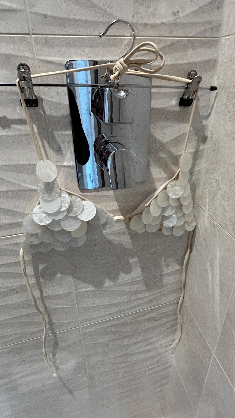 Puede incluir: Un top de bikini de conchas blancas colgado de una percha. El top de bikini est&aacute; hecho de peque&ntilde;as conchas blancas redondas y tiene finas correas beige. El fondo incluye un grifo de ducha cromado y baldosas de color gris claro.