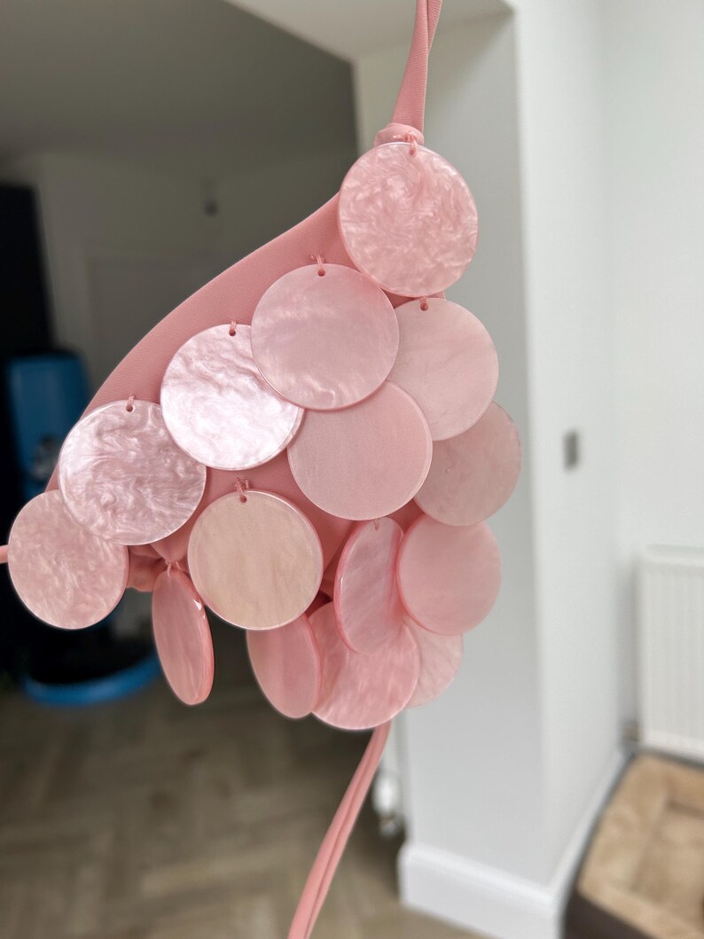 Puede incluir: Top de bikini rosa claro con dise&ntilde;o de cuello halter. El top est&aacute; adornado con m&uacute;ltiples discos redondos, iridiscentes y rosas. Los discos est&aacute;n sujetos al top con peque&ntilde;os lazos. El top de bikini est&aacute; hecho de un material suave y liso.