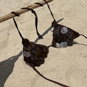 Puede incluir: Un top de bikini marr&oacute;n hecho de peque&ntilde;as piezas redondas de color marr&oacute;n oscuro y plateado. El top de bikini est&aacute; suspendido de un palo de madera con lazos marrones. El fondo es una playa de arena.