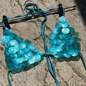 May include: A turquoise bikini top with a unique design. The triangle cups are covered in overlapping, iridescent, circular sequins. The straps are a matching turquoise colour. The bikini is displayed on a hanger.
