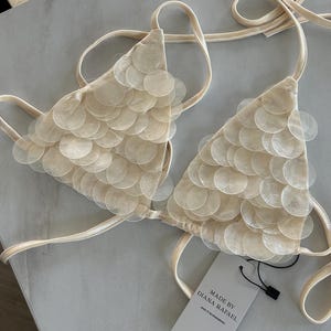 May include: A cream-colored bikini top featuring two triangular cups adorned with overlapping, translucent circular sequins. The garment includes thin tie straps and a white rectangular tag with the text MADE BY DIANA RAFAEL.