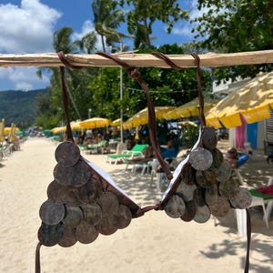 Puede incluir: Un top de bikini marr&oacute;n adornado con decoraciones circulares. La parte superior est&aacute; hecha de tela marr&oacute;n y presenta numerosos discos peque&ntilde;os, redondos y con efecto met&aacute;lico. Las correas tambi&eacute;n son marrones. El fondo muestra una escena de playa con sombrillas y personas.