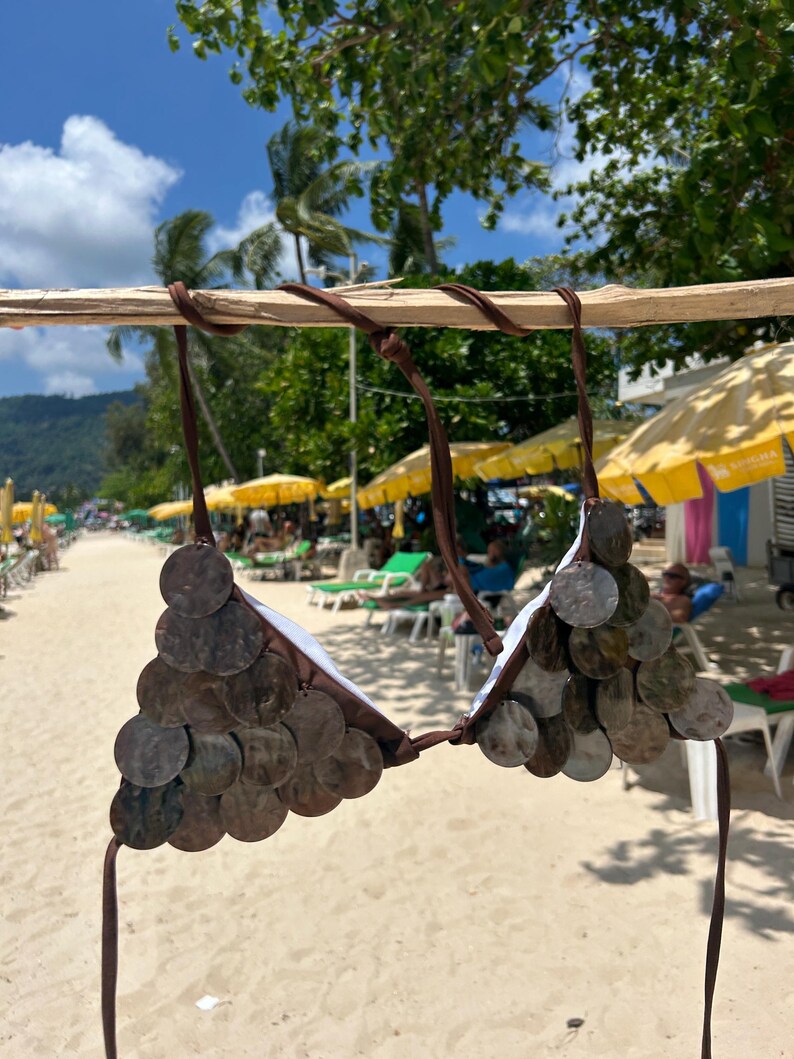 Puede incluir: Un top de bikini marr&oacute;n adornado con decoraciones circulares. La parte superior est&aacute; hecha de tela marr&oacute;n y presenta numerosos discos peque&ntilde;os, redondos y con efecto met&aacute;lico. Las correas tambi&eacute;n son marrones. El fondo muestra una escena de playa con sombrillas y personas.