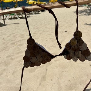 Puede incluir: Un top de bikini marr&oacute;n hecho de monedas, exhibido en un palo de madera contra un fondo de playa arenosa. El top de bikini presenta copas cubiertas de monedas y correas marrones. Se ven sombrillas amarillas y sillas de playa en el fondo.