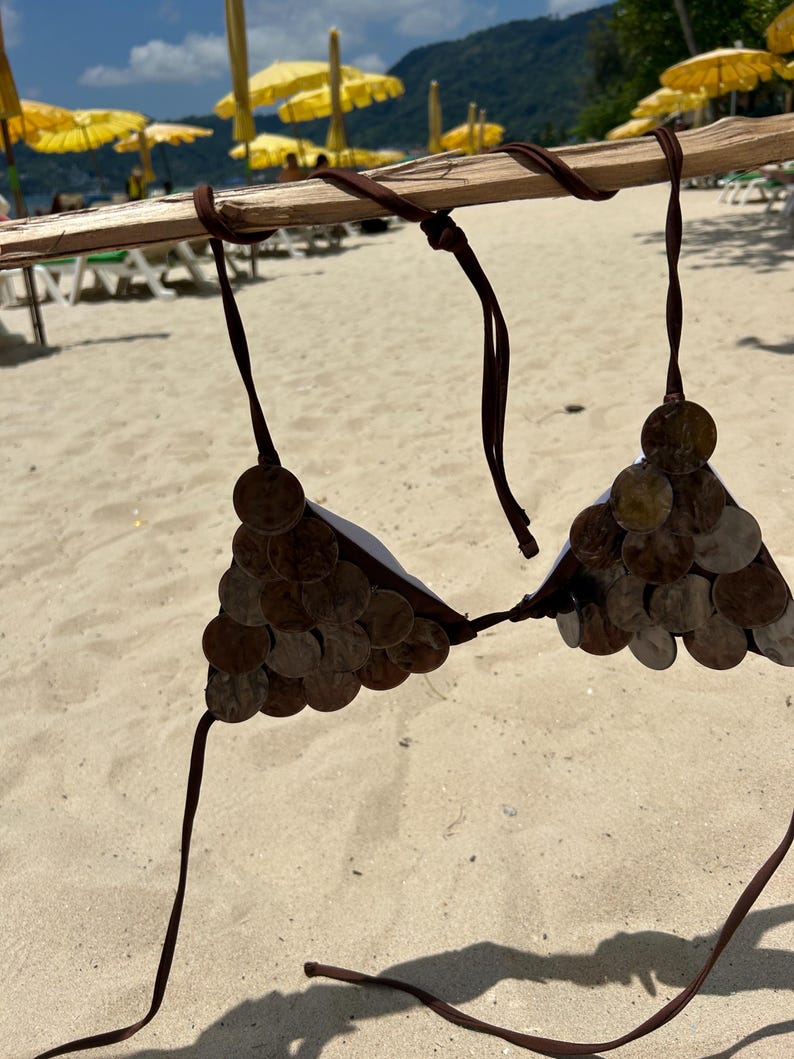 Puede incluir: Un top de bikini marr&oacute;n hecho de monedas, exhibido en un palo de madera contra un fondo de playa arenosa. El top de bikini presenta copas cubiertas de monedas y correas marrones. Se ven sombrillas amarillas y sillas de playa en el fondo.