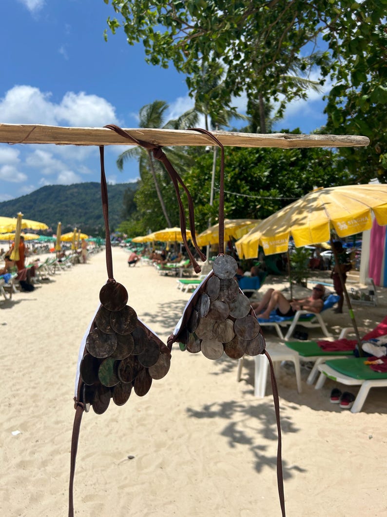 Puede incluir: Un top de bikini &uacute;nico hecho de peque&ntilde;as piezas met&aacute;licas redondas y oscuras, posiblemente monedas, adheridas a una tela blanca. El bikini cuelga de una barra de madera, con correas marrones. El fondo muestra una escena de playa con sombrillas amarillas y cielo azul.