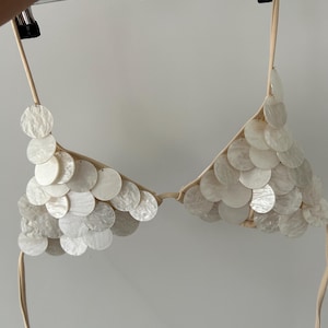 Puede incluir: Un top de bikini blanco adornado con numerosas lentejuelas redondas e iridiscentes. El top est&aacute; suspendido de una percha, mostrando su forma triangular y sus delicadas correas. Las lentejuelas crean un efecto brillante, sugiriendo un tema de playa o traje de ba&ntilde;o.