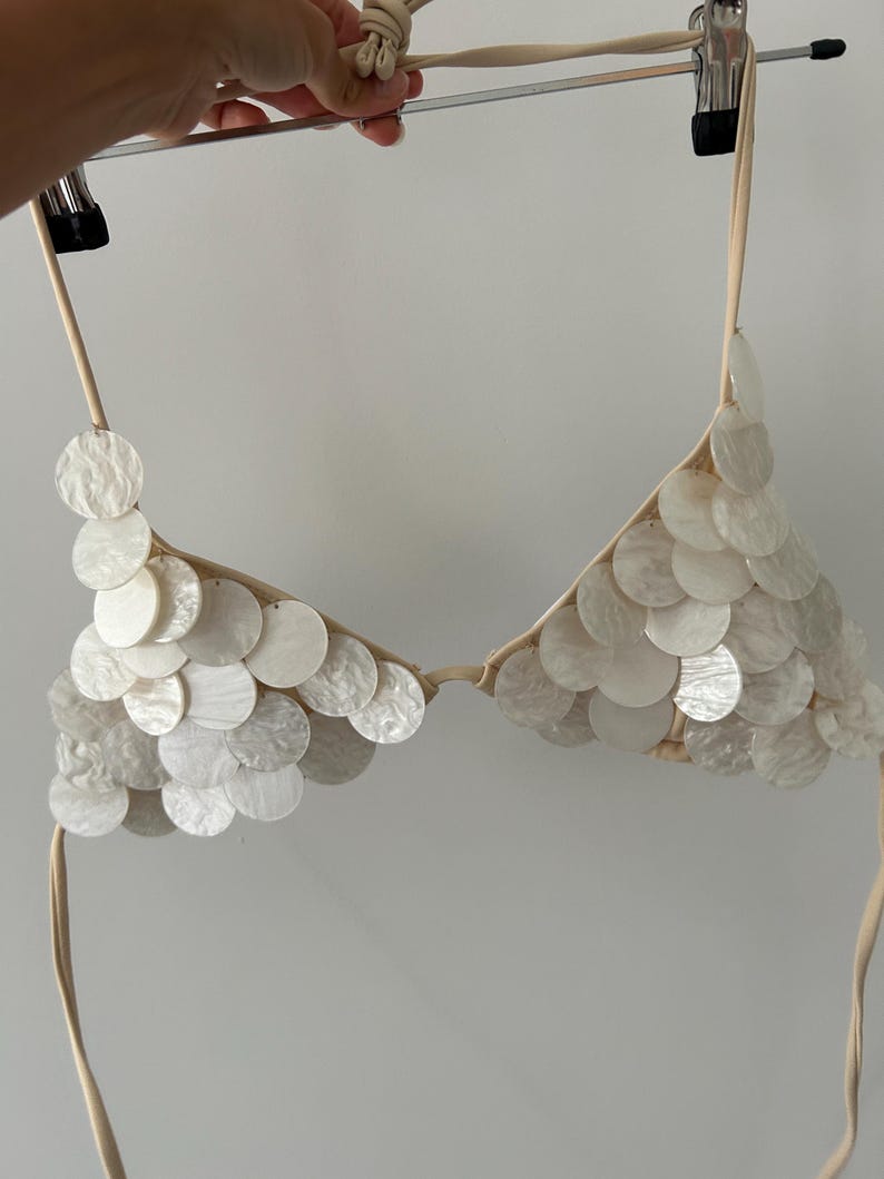 Puede incluir: Un top de bikini blanco adornado con numerosas lentejuelas redondas e iridiscentes. El top est&aacute; suspendido de una percha, mostrando su forma triangular y sus delicadas correas. Las lentejuelas crean un efecto brillante, sugiriendo un tema de playa o traje de ba&ntilde;o.
