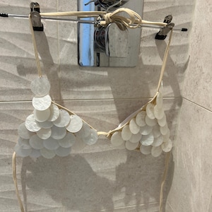 Puede incluir: Un top de bikini blanco adornado con numerosas piezas redondas e iridiscentes de concha. El top est&aacute; suspendido de una percha, mostrando su delicado dise&ntilde;o y materiales naturales. El top de bikini est&aacute; colgado en una barra de metal frente a una pared alicatada.