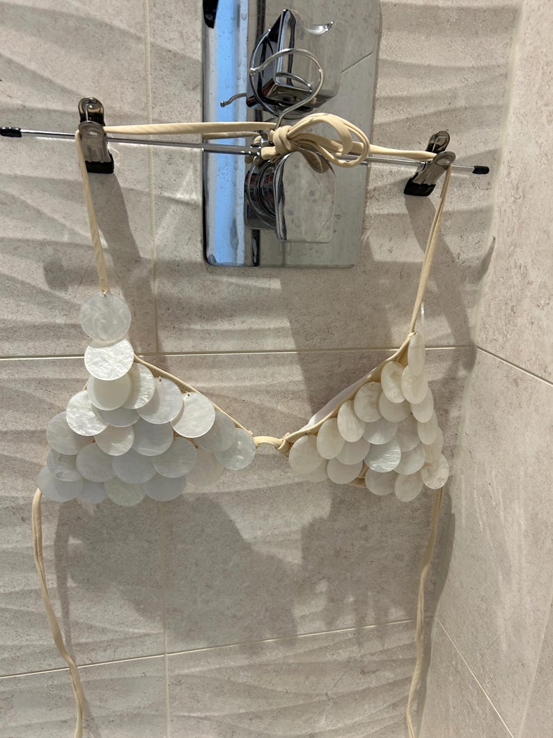 Puede incluir: Un top de bikini blanco adornado con numerosas piezas redondas e iridiscentes de concha. El top est&aacute; suspendido de una percha, mostrando su delicado dise&ntilde;o y materiales naturales. El top de bikini est&aacute; colgado en una barra de metal frente a una pared alicatada.