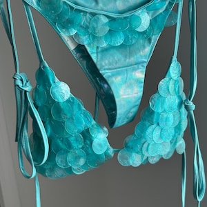 May include: A turquoise bikini set featuring a shimmering, iridescent fabric base. The triangle top and bikini bottoms are heavily embellished with overlapping, translucent circular sequins that create a textured, scale-like appearance throughout.