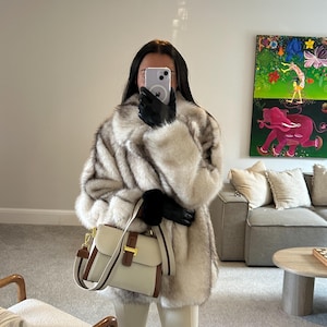 May include: A fluffy, long-sleeved, cream and gray faux fur coat paired with cream-colored trousers and black gloves. A structured, cream-colored leather handbag with brown leather trim and a gold-toned metal clasp is held in the hand.
