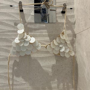 Puede incluir: Un top de bikini color crema con un dise&ntilde;o &uacute;nico. Est&aacute; adornado con numerosos adornos circulares y superpuestos en forma de concha. El top de bikini est&aacute; suspendido de una percha, mostrando sus intrincados detalles y textura.