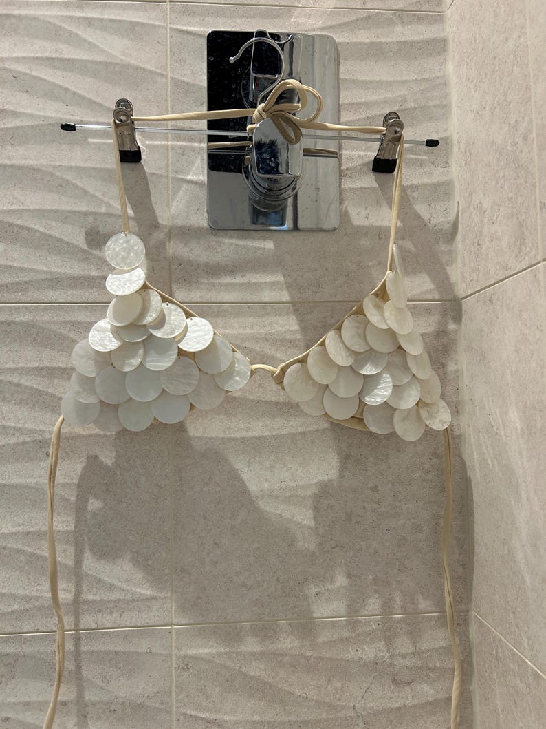 Puede incluir: Un top de bikini color crema con un dise&ntilde;o &uacute;nico. Est&aacute; adornado con numerosos adornos circulares y superpuestos en forma de concha. El top de bikini est&aacute; suspendido de una percha, mostrando sus intrincados detalles y textura.