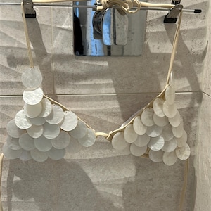May include: White bikini top with circular shell-like embellishments. The top is suspended from a clothes hanger. The bikini top is made of a light-colored material and has thin straps for tying. The background is a tiled wall.