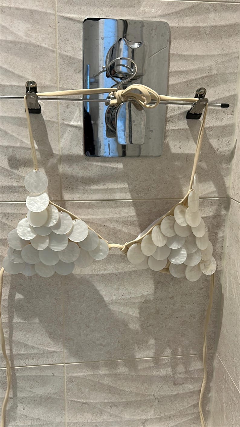 Puede incluir: Top de bikini blanco con adornos circulares en forma de concha. El top est&aacute; suspendido de una percha. El top de bikini est&aacute; hecho de un material de color claro y tiene finas correas para atar. El fondo es una pared alicatada.