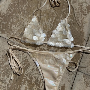 May include: A cream-colored bikini set. The triangle top and the bikini bottom are adorned with iridescent, circular embellishments. The bikini top has halter straps, and the bottom has tie sides. The fabric has a subtle sheen.