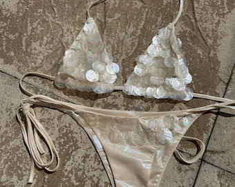 May include: A cream-colored bikini set. The triangle top and the bikini bottom are adorned with iridescent, circular embellishments. The bikini top has halter straps, and the bottom has tie sides. The fabric has a subtle sheen.