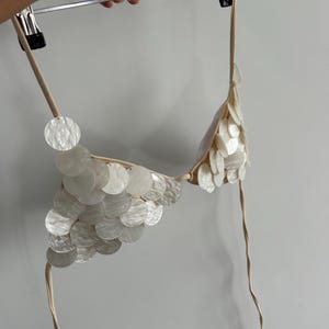 Puede incluir: Un top de bikini color crema adornado con numerosos adornos circulares iridiscentes. El top de bikini est&aacute; suspendido de una percha, mostrando su delicado dise&ntilde;o y el brillo sutil de los elementos decorativos. Las correas son de color crema a juego.