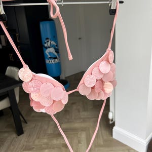 Puede incluir: Top de bikini rosa claro con lentejuelas circulares. El top de bikini est&aacute; colgado de una percha plateada. Las lentejuelas son de diferentes tonos de rosa. El bikini est&aacute; atado con finas correas rosas.