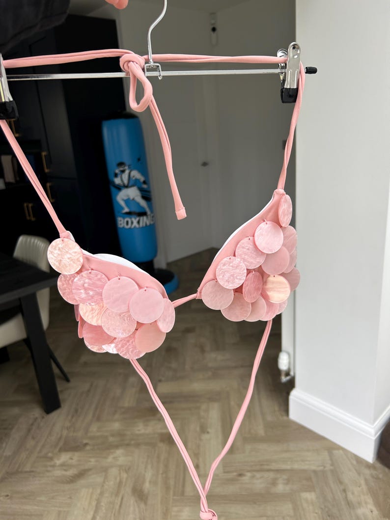 Puede incluir: Top de bikini rosa claro con lentejuelas circulares. El top de bikini est&aacute; colgado de una percha plateada. Las lentejuelas son de diferentes tonos de rosa. El bikini est&aacute; atado con finas correas rosas.