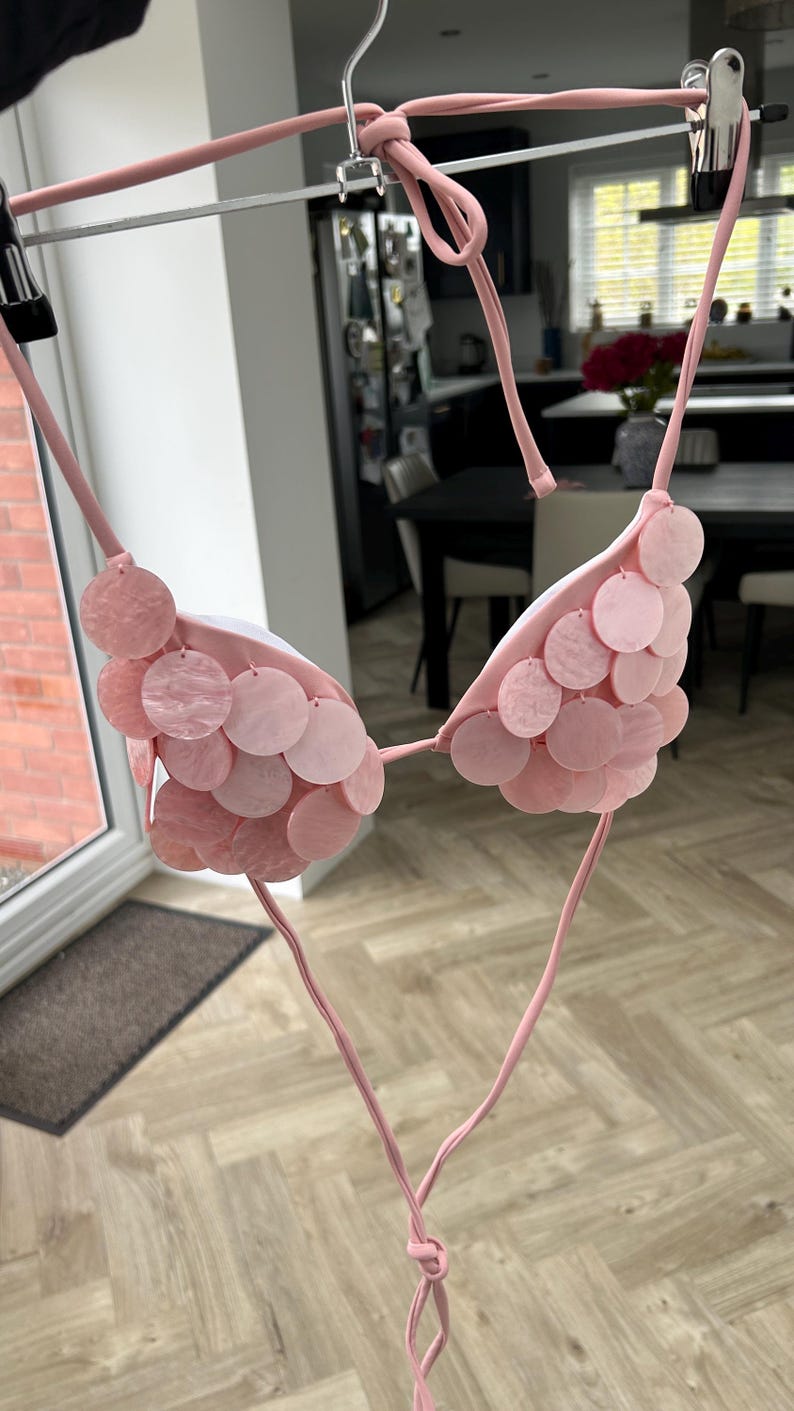 Puede incluir: Top de bikini rosa claro con adornos circulares. El top est&aacute; suspendido de una percha. Presenta un dise&ntilde;o de cuello halter y lazos. Los adornos circulares son de diferentes tonos de rosa.