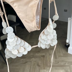May include: A beige bikini set. The top is decorated with white, circular embellishments. The bottoms are a solid beige colour. The bikini is hanging on a hanger.