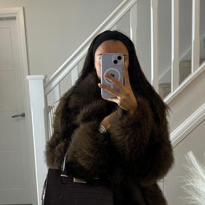 Può includere: Un cappotto in pelliccia sintetica marrone scuro con una consistenza spessa e morbida e maniche lunghe. Il cappotto è abbinato a una borsa strutturata bordeaux scuro con texture effetto coccodrillo, dettagli dorati e un manico superiore.
