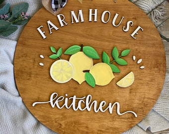 May include: A round wooden farmhouse kitchen sign with the words "FARMHOUSE kitchen" in white script. The sign features a lemon and leaf design with a buffalo check bow.