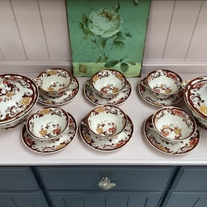 Może przedstawiać: Zestaw do herbaty w stylu vintage z białymi porcelanowymi filiżankami, spodkami i naczyniami do serwowania. Zestaw posiada kwiatowy wzór w kolorze czerwonym, żółtym i brązowym. Zawiera zakryte miski do serwowania i wiele filiżanek i spodków.