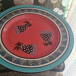 Pode incluir: Um prato grande, redondo e decorativo com uma borda turquesa e um centro vermelho. O prato é decorado com um padrão geométrico em preto, branco e dourado. O centro do prato apresenta três penas pretas com pontos brancos sobre um fundo vermelho.