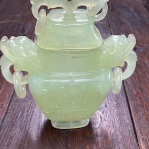 May include: A pale green jade-like decorative jar with a lid, intricate carvings, and ring handles. The jar has a rounded body with floral designs and is set on a dark wooden surface. The lid is topped with a decorative floral design.
