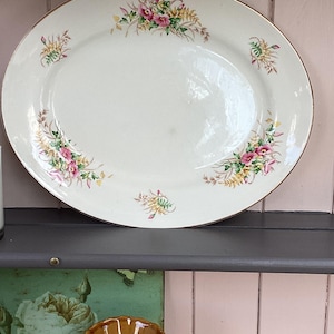 Peut inclure: Un plat de service ovale en céramique avec une base de couleur crème et un fin bord doré. Le plat présente des motifs floraux délicats dans des tons de rose, jaune et vert, disposés en petits bouquets autour du bord de la surface.