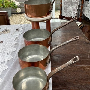 May include: Four copper saucepans with long handles, nested together on a wooden table. The saucepans are shiny and have a slight patina.