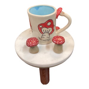 May include: A white ceramic mug with a blue interior and red and white mushroom illustrations. The mug is sitting on a white marble table with a wooden leg and two red and white mushroom figurines.