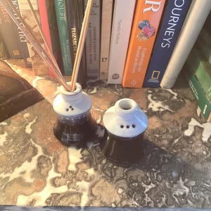 Pode incluir: Duas garrafas difusoras de cana de cerâmica com tampo branco e base castanha escura. Uma garrafa tem canas inseridas. As garrafas estão sobre uma superfície de mármore, com um pano de fundo de livros coloridos.