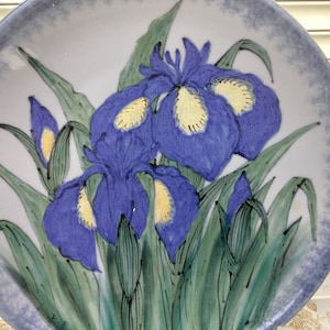May include: A white ceramic plate with a blue and yellow floral design. The plate features a cluster of irises with green leaves and stems. The background is a light blue with a white centre.