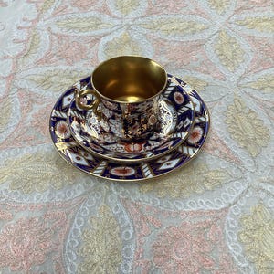 Può includere: Un set di tazza da tè e piattino in porcellana caratterizzato da un motivo floreale blu scuro e bianco con intricate finiture dorate. L'interno della tazza è rifinito in oro metallizzato, e anche il manico è dorato, abbinato ai bordi del piattino.
