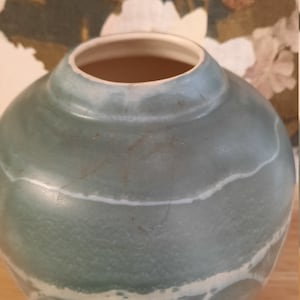 May include: A round, pale blue ceramic vase with a wide opening. The vase has a textured surface and is decorated with white, irregular lines. The interior is a light brown colour. The vase is on a wooden surface.