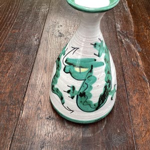 Könnte beinhalten: Eine Keramikvase mit weißem Körper und grünen Akzenten. Die Vase hat einen ausgestellten oberen Rand und einen bauchigen Boden. Grüne abstrakte Designs verzieren den Körper der Vase. Die Vase steht auf einer Holzoberfläche.