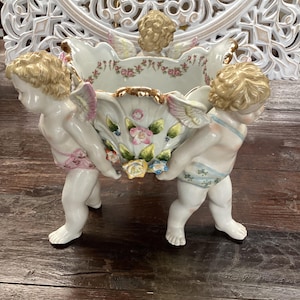 Puede incluir: Un macetero de porcelana blanca con tres querubines sosteniendo el macetero. El macetero está decorado con flores rosas, amarillas y azules y ribetes dorados.