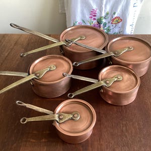 May include: A collection of copper pots with brass handles and lids. The pots are arranged on a wooden surface, showcasing their shiny, reflective surfaces. The handles are riveted to the pots, and the lids fit snugly. The overall aesthetic is vintage and classic.