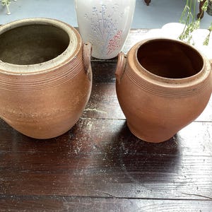 May include: Two antique, brown ceramic pots with handles. The pots have a wide opening and a textured, aged appearance. The pots are on a dark wooden surface. A white vase is in the background.