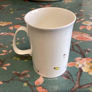 Może przedstawiać: Biały ceramiczny kubek z zakrzywionym uchwytem. Kubek ma małe ilustracje biedronek i innych owadów. Kubek stoi na powierzchni z kwiatowym wzorem.