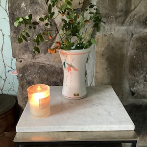 Op de afbeelding: Een witte keramische vaas met een uitlopende rand en een abstract geschilderd ontwerp in koraal en blauwgroen, met een groene tak met kleine oranje bessen. Naast de vaas staat een cilindrische glazen kaarsenhouder met een brandende witte kaars.